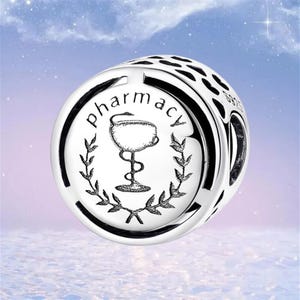 May include: A silver charm with the word "pharmacy" encircling a caduceus symbol. The charm has a detailed design with a black border and a series of small cutouts. The background is a soft gradient of blue and purple.