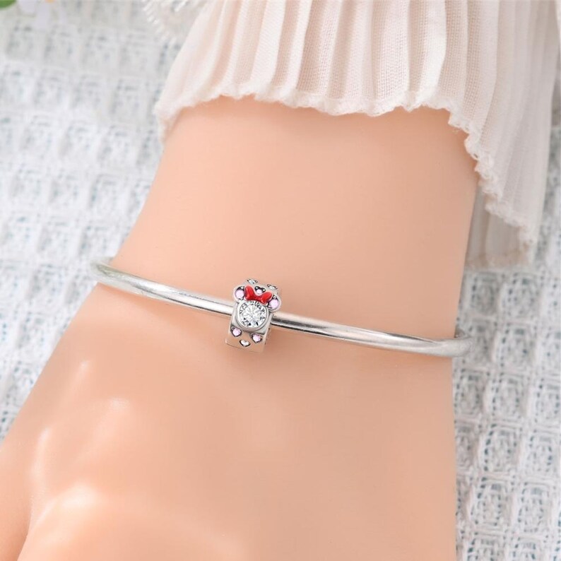 K&ouml;nnte beinhalten: Ein silberfarbenes Armband mit einem einzelnen Anh&auml;nger. Der Anh&auml;nger ist in Form eines Mauskopfes mit roter Schleife gestaltet und mit kleinen rosa Steinen sowie einem zentralen, klaren runden Stein besetzt. Das Armband hat eine glatte Oberfl&auml;che.