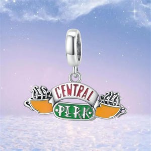 May include: A silver charm with the "Central Perk" logo from the TV show Friends. The charm features a banner with the words "Central Perk" in red and white, and a green oval with the words "Perk" in white. Two orange coffee cups are on either side.