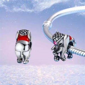 May include: Two silver elephant charms with red accents and a pink gem eye. One charm is attached to a silver bracelet. The charms have detailed textures and are set against a soft, blue background.