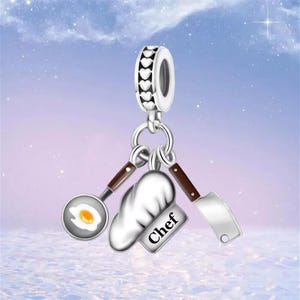 May include: A silver charm with a chef theme. The charm features a frying pan with a fried egg, a chef's hat, and a cleaver. The charm also has a silver ring with heart-shaped cutouts.