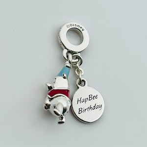 May include: A silver charm with a Winnie the Pooh figure wearing a red vest and blue party hat. A round charm with the words "HapBee Birthday" hangs below. The charm is marked with the Disney logo.