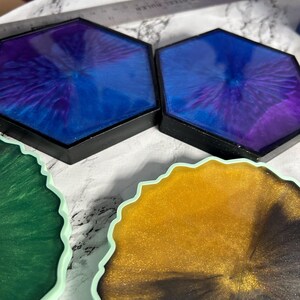 May include: A collection of handmade resin coasters in various colors. Two hexagonal coasters feature a deep blue and purple swirl design with black edges. Other coasters have a green, gold, and black design with a scalloped edge.