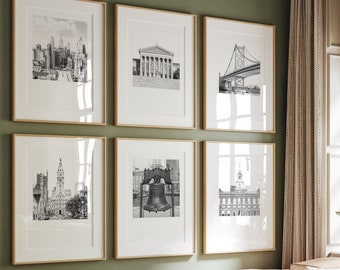 May include: A living room with a green wall and a light brown leather couch. There are six black and white framed photos on the wall, each depicting a different landmark in Philadelphia. The photos are arranged in a grid pattern. The couch has a green throw pillow and a striped throw blanket. There is a small table in front of the couch with a book and a vase of white flowers.