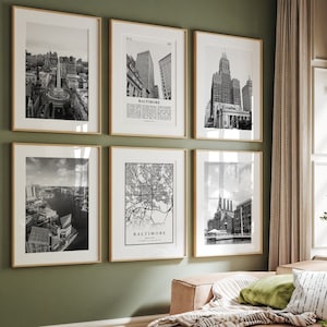 May include: A living room with a green wall and a light brown sofa. There are six black and white framed photographs on the wall, featuring cityscapes and a map of Baltimore. The photographs are arranged in a grid pattern. The sofa has a green throw pillow and a striped throw blanket. There is a small table in front of the sofa with a book and a vase of flowers on it.