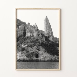 May include: Black and white photograph of a rocky cliff face with a tall, pointed rock formation rising from the center. The cliff face is covered in vegetation and there is a small cave opening near the base. The water is calm and reflects the sky.
