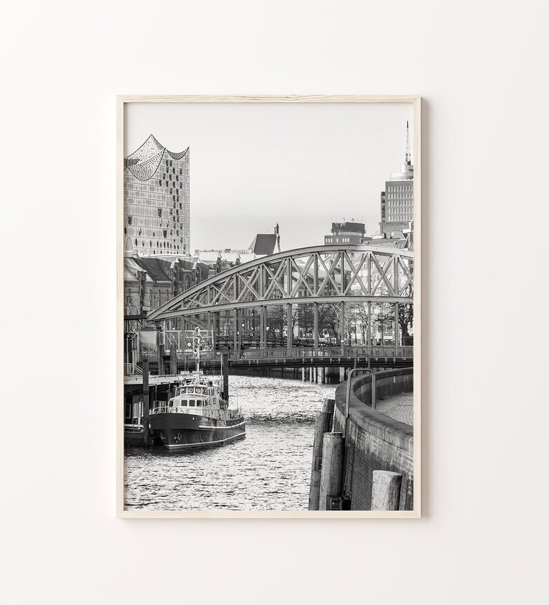 May include: A black and white photograph of a city skyline with a bridge over a river. The bridge is made of metal and has a truss design. There is a boat in the river.
