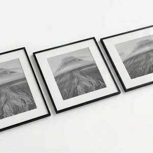 May include: Three framed black and white landscape prints. Each frame has a black border and a white mat. The prints depict a mountain range with a cloudy sky. The artwork is displayed on a white surface.
