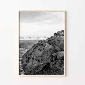 May include: Black and white photograph of a rocky mountain peak with a group of people standing on the top. The mountain is in the foreground and the background is a valley with rolling hills and clouds.