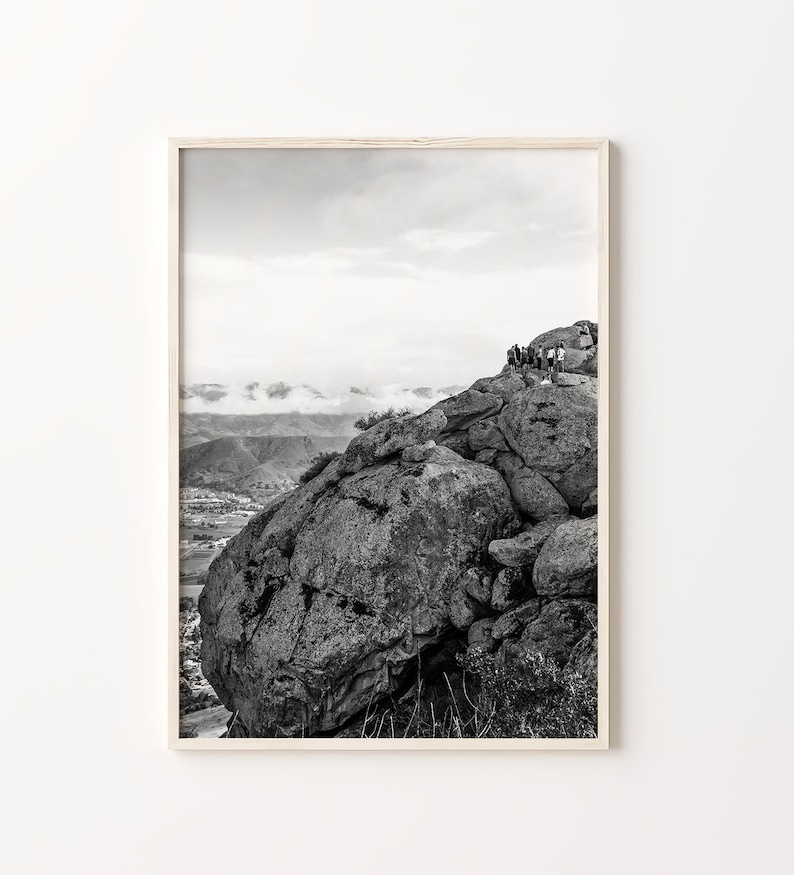 May include: Black and white photograph of a rocky mountain peak with a group of people standing on the top. The mountain is in the foreground and the background is a valley with rolling hills and clouds.