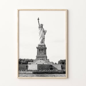May include: Black and white framed print of the Statue of Liberty. The iconic monument stands tall against a bright sky, with the base and surrounding water visible. The print is in a light-coloured wooden frame.