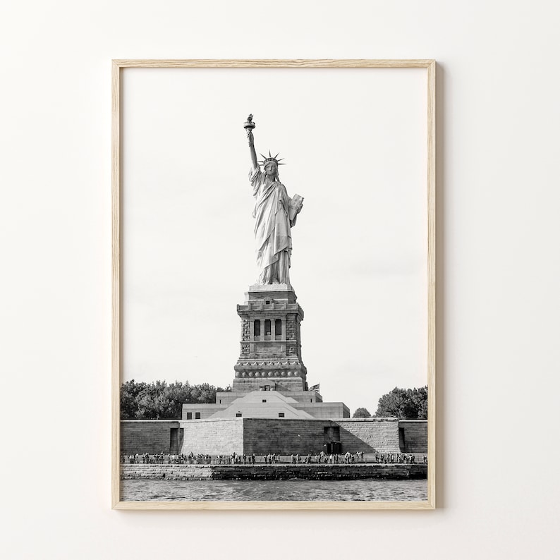 May include: Black and white framed print of the Statue of Liberty. The iconic monument stands tall against a bright sky, with the base and surrounding water visible. The print is in a light-coloured wooden frame.