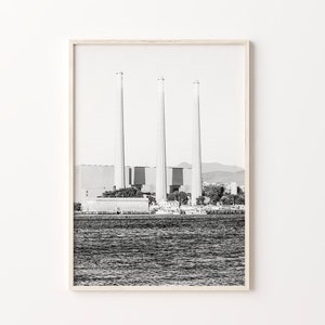 May include: Black and white photograph of three tall smokestacks against a cloudy sky. The smokestacks are part of a large industrial complex, which is located on a body of water. The water is calm and reflects the sky.