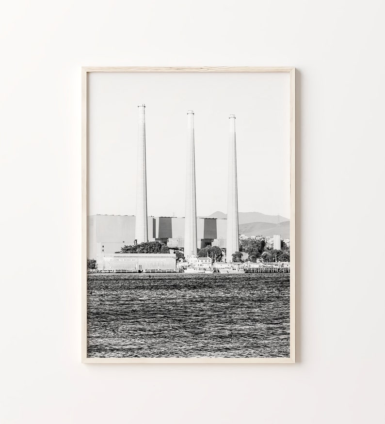 May include: Black and white photograph of three tall smokestacks against a cloudy sky. The smokestacks are part of a large industrial complex, which is located on a body of water. The water is calm and reflects the sky.