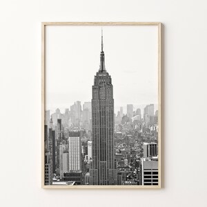 May include: Monochrome framed print of the Empire State Building, a famous New York City landmark. The image shows the skyscraper against a backdrop of other buildings. The frame is made of light-coloured wood.