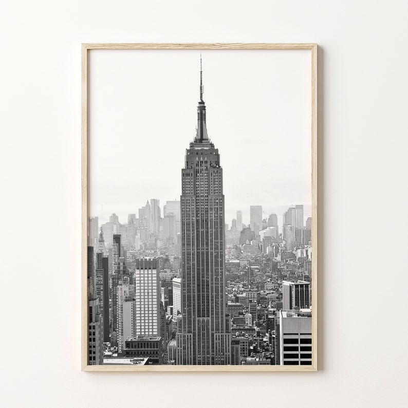 May include: Monochrome framed print of the Empire State Building, a famous New York City landmark. The image shows the skyscraper against a backdrop of other buildings. The frame is made of light-coloured wood.