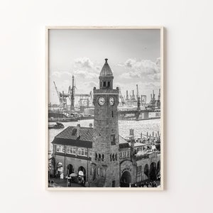 May include: A black and white photograph of a clock tower building with a clock face on the front. The building is located on a waterfront with a harbor in the background. There are cranes and ships in the harbor.