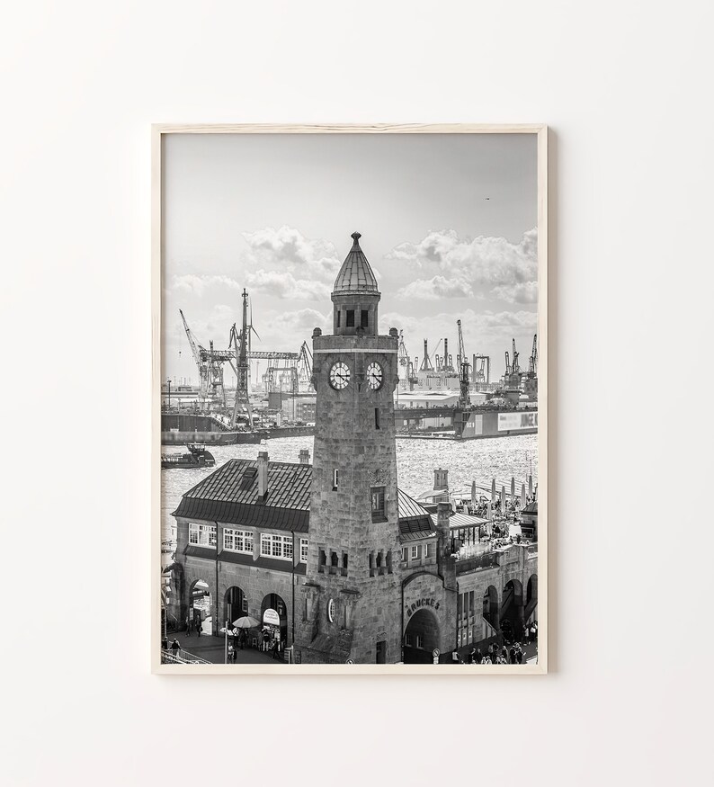 May include: A black and white photograph of a clock tower building with a clock face on the front. The building is located on a waterfront with a harbor in the background. There are cranes and ships in the harbor.
