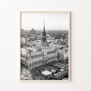 May include: Black and white aerial photograph of a city with a tall building in the center. The building has a clock tower and a spire. There are many smaller buildings surrounding the central building. There are people walking in the streets below.