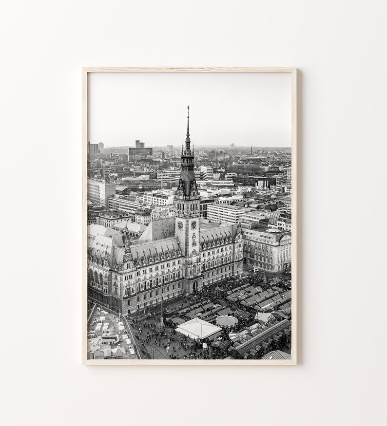 May include: Black and white aerial photograph of a city with a tall building in the center. The building has a clock tower and a spire. There are many smaller buildings surrounding the central building. There are people walking in the streets below.