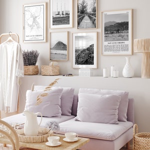 May include: A living room interior with a light purple sofa, a wooden coffee table, and a white pitcher on a woven tray. The wall behind the sofa is decorated with black and white framed prints of maps, landscapes, and cityscapes. The prints include the text "San Luis Obispo, California".