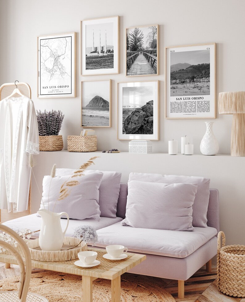 May include: A living room interior with a light purple sofa, a wooden coffee table, and a white pitcher on a woven tray. The wall behind the sofa is decorated with black and white framed prints of maps, landscapes, and cityscapes. The prints include the text "San Luis Obispo, California".