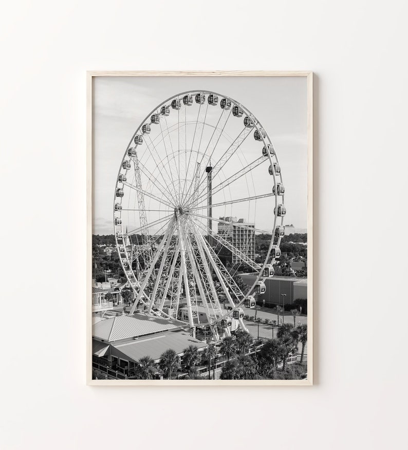 May include: A black and white photograph of a Ferris wheel at an amusement park. The Ferris wheel is in the center of the image and is surrounded by buildings and trees.