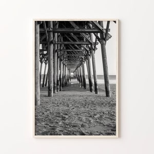 May include: Black and white photograph of a wooden pier extending over a sandy beach. The image is taken from underneath the pier, looking towards the end of the pier.