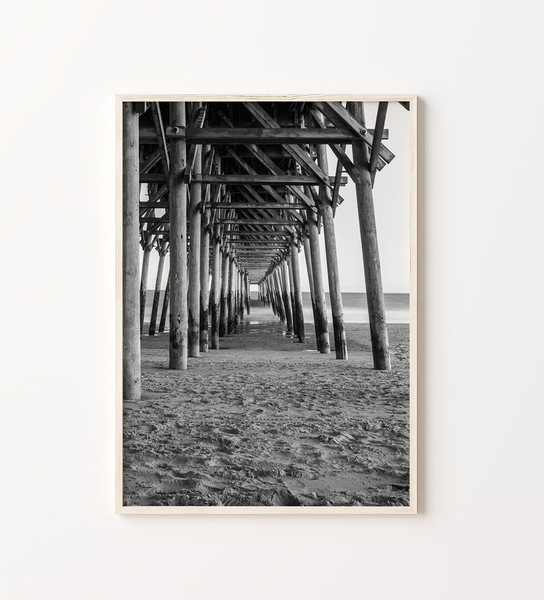 May include: Black and white photograph of a wooden pier extending over a sandy beach. The image is taken from underneath the pier, looking towards the end of the pier.