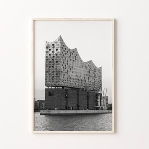 May include: A black and white photograph of a modern building with a unique, angular design. The building is situated on a waterfront with cranes in the background.