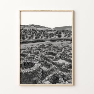 May include: Black and white photograph of ancient ruins in a field. The ruins are made of stone and are surrounded by trees and hills.