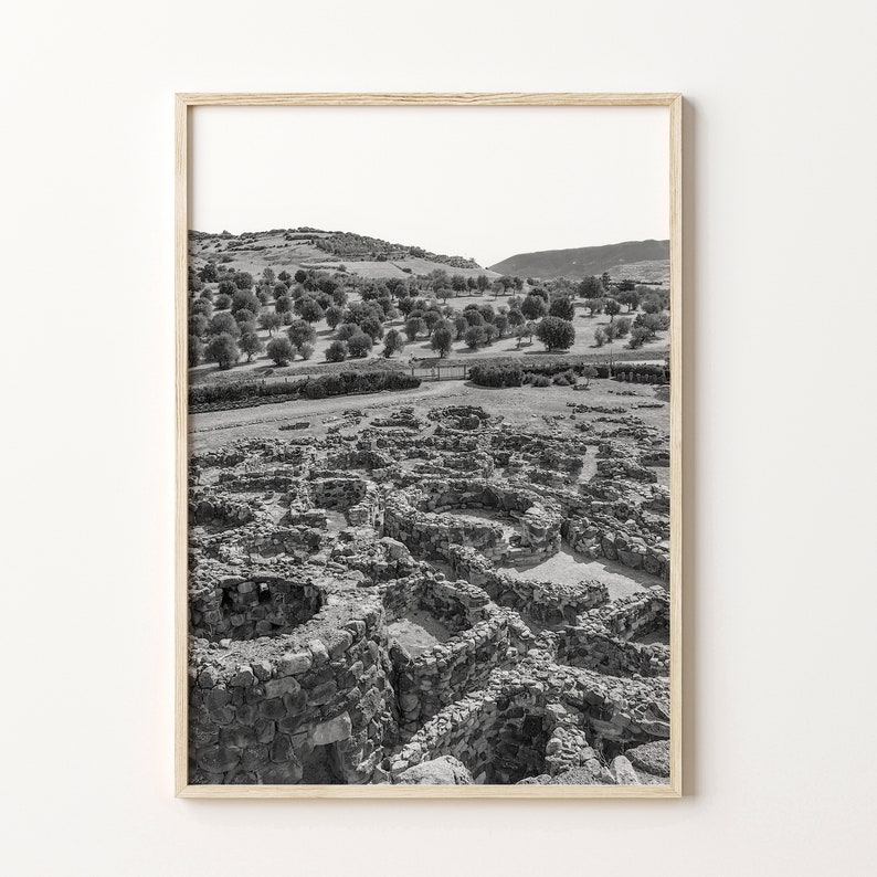 May include: Black and white photograph of ancient ruins in a field. The ruins are made of stone and are surrounded by trees and hills.