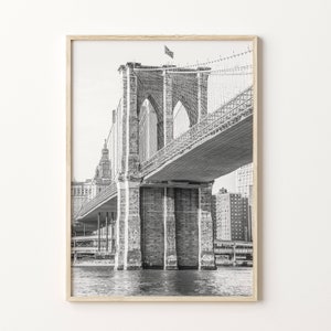 May include: Black and white photograph of the Brooklyn Bridge, framed in light wood. The bridge's stone towers and suspension cables are visible. An American flag flies atop one tower. The water is visible below the bridge.