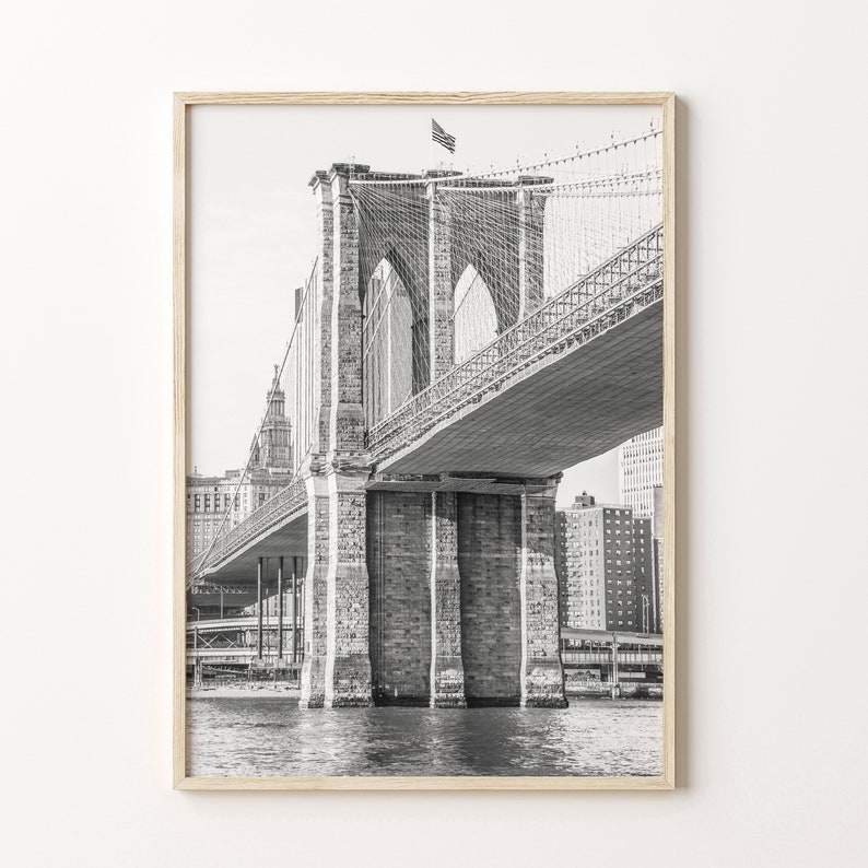 May include: Black and white photograph of the Brooklyn Bridge, framed in light wood. The bridge's stone towers and suspension cables are visible. An American flag flies atop one tower. The water is visible below the bridge.