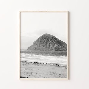May include: A black and white photograph of a rocky island in the ocean. The island is in the distance, and the foreground shows a sandy beach with waves crashing on the shore.