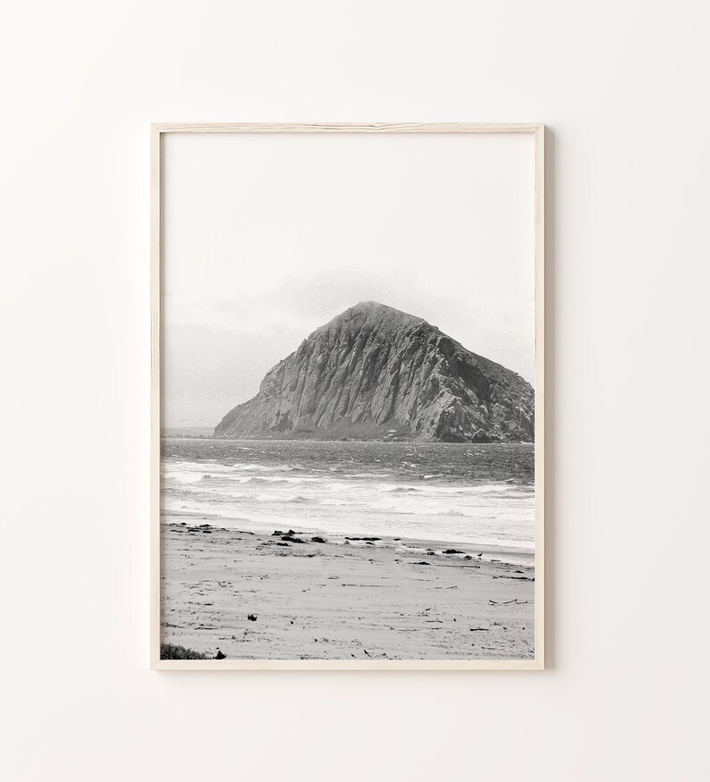 May include: A black and white photograph of a rocky island in the ocean. The island is in the distance, and the foreground shows a sandy beach with waves crashing on the shore.