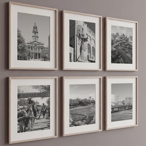 May include: Six framed black and white photographs with light wood frames. The images depict cityscapes, architecture, and a scene with longhorn cattle and the text "STOCKYARDS STATION". The frames are arranged on a neutral-coloured wall.