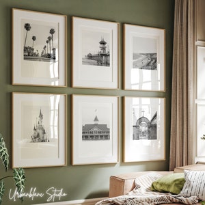 Può includere: Una parete di galleria con sei fotografie in bianco e nero incorniciate. Le foto mostrano varie scene, tra cui una spiaggia con palme, un molo, un castello e un edificio con un campanile.