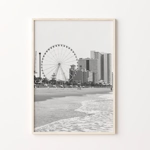 May include: Black and white photograph of a Ferris wheel on a beach with buildings in the background.