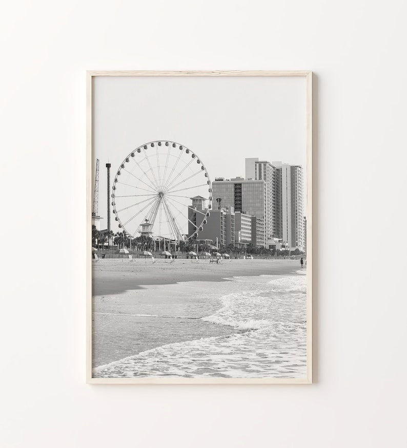 May include: Black and white photograph of a Ferris wheel on a beach with buildings in the background.