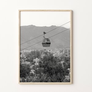 Puede incluir: Una fotograf&iacute;a en blanco y negro de un telef&eacute;rico que viaja sobre una ciudad con monta&ntilde;as en el fondo.