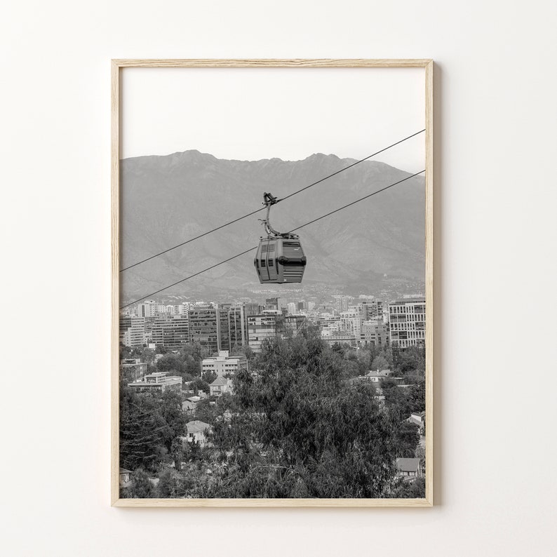 Puede incluir: Una fotograf&iacute;a en blanco y negro de un telef&eacute;rico que viaja sobre una ciudad con monta&ntilde;as en el fondo.