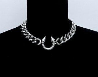 May include: A silver-toned metal chain necklace featuring a thick curb link design. The center of the necklace showcases a U-shaped horseshoe-style pendant with two pointed, cone-shaped spikes extending upward from each end of the curve.