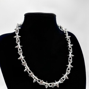 以下が含まれることがあります： 尖ったスパイクが付いたシルバーチェーンネックレス。ネックレスは黒いマネキンバストの上にあります。