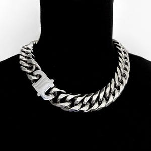 Può includere: Una collana a catena argentata e massiccia con una grande chiusura. La collana è composta da maglie spesse e interconnesse ed è mostrata su uno sfondo nero. La chiusura è una caratteristica importante, che si aggiunge al design audace della collana.