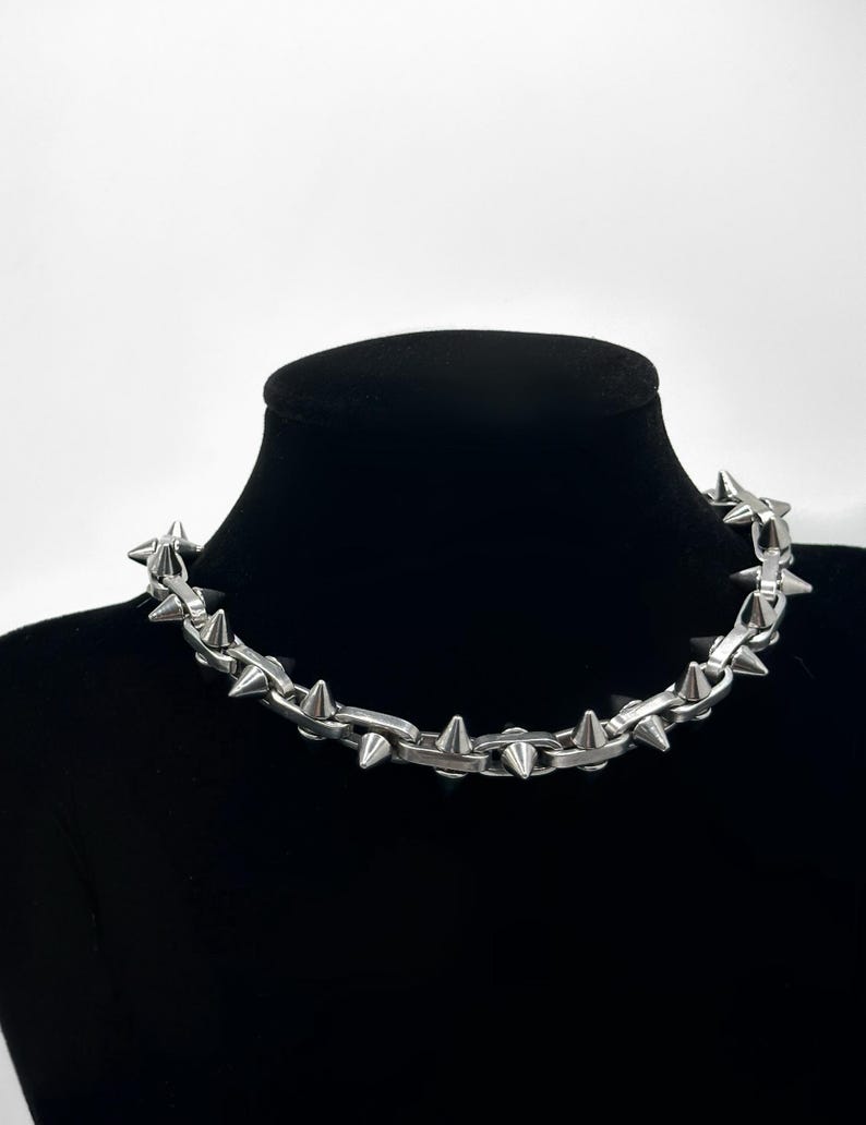 May include: A silver chain necklace with pointed spikes. The necklace is displayed on a black velvet mannequin bust.
