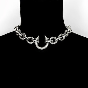 Puede incluir: Un collar gargantilla plateado con un diseño audaz y vanguardista. El collar presenta un estilo de eslabones de cadena con un elemento central en forma de herradura. Dos puntas cónicas y afiladas están posicionadas en la herradura.