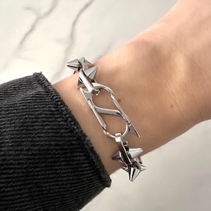 May include: A silver-colored bracelet featuring a chain of spiked, star-shaped links. The bracelet has a unique design with a central, stylized clasp. The jewelry is worn on a wrist, showcasing its edgy and modern aesthetic.