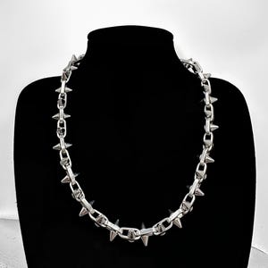 以下が含まれることがあります： 尖ったスパイクが付いたシルバーチェーンネックレス。ネックレスは黒いマネキンに着けられています。