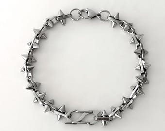 May include: A silver-toned metal chain bracelet featuring a series of evenly spaced, pointed conical spikes along the links. The bracelet is secured with a lobster claw clasp and includes a distinct, angular geometric link component.
